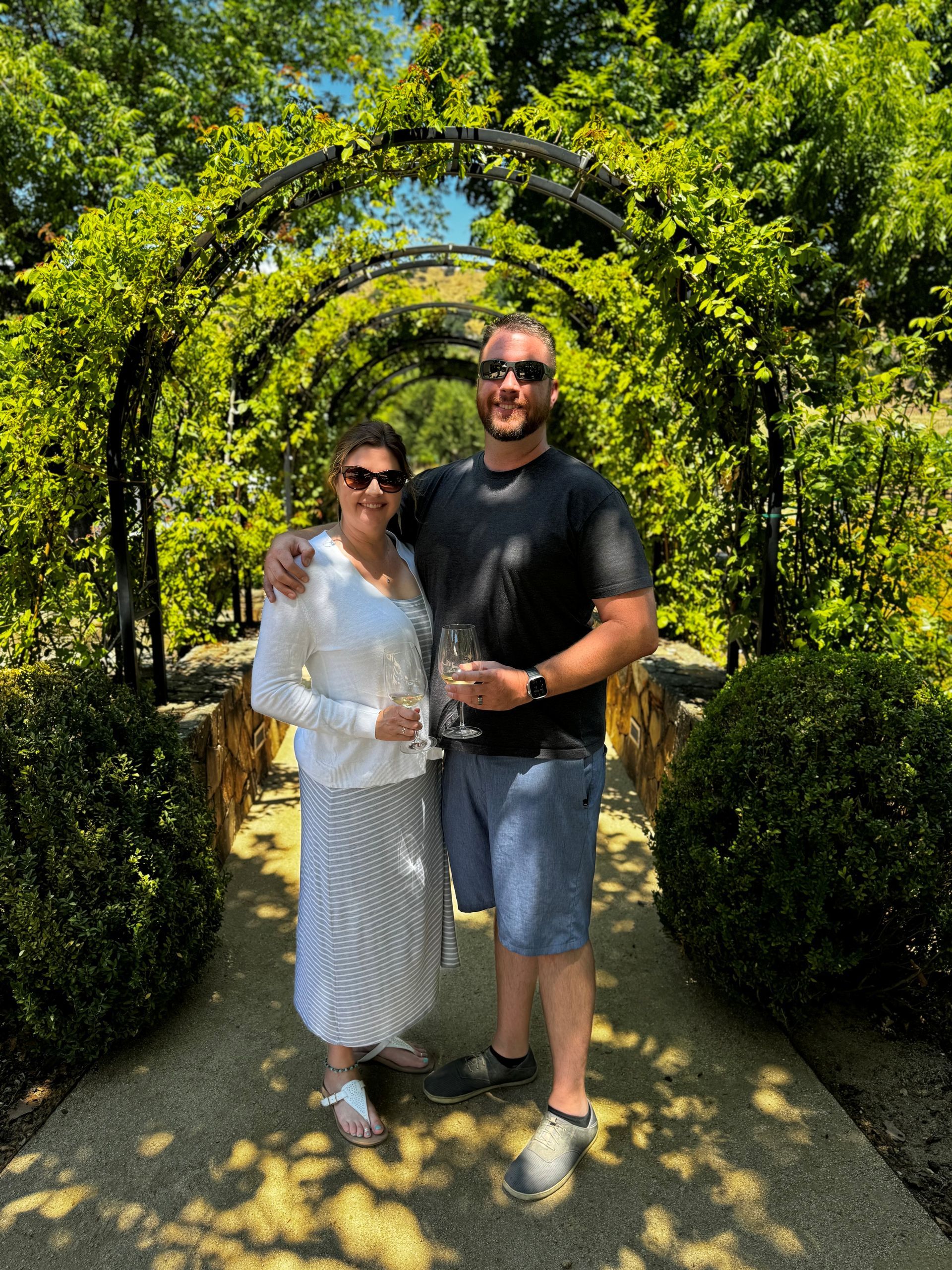 Couple standing under a vine-covered archway on a sunny garden path, holding drinks and smiling.