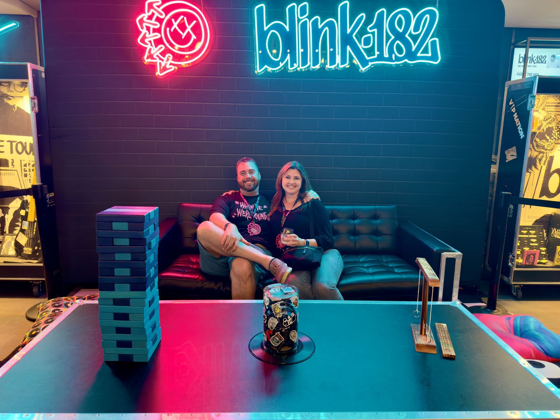 Two people on a couch under a neon Blink-182 sign in a dimly lit lounge