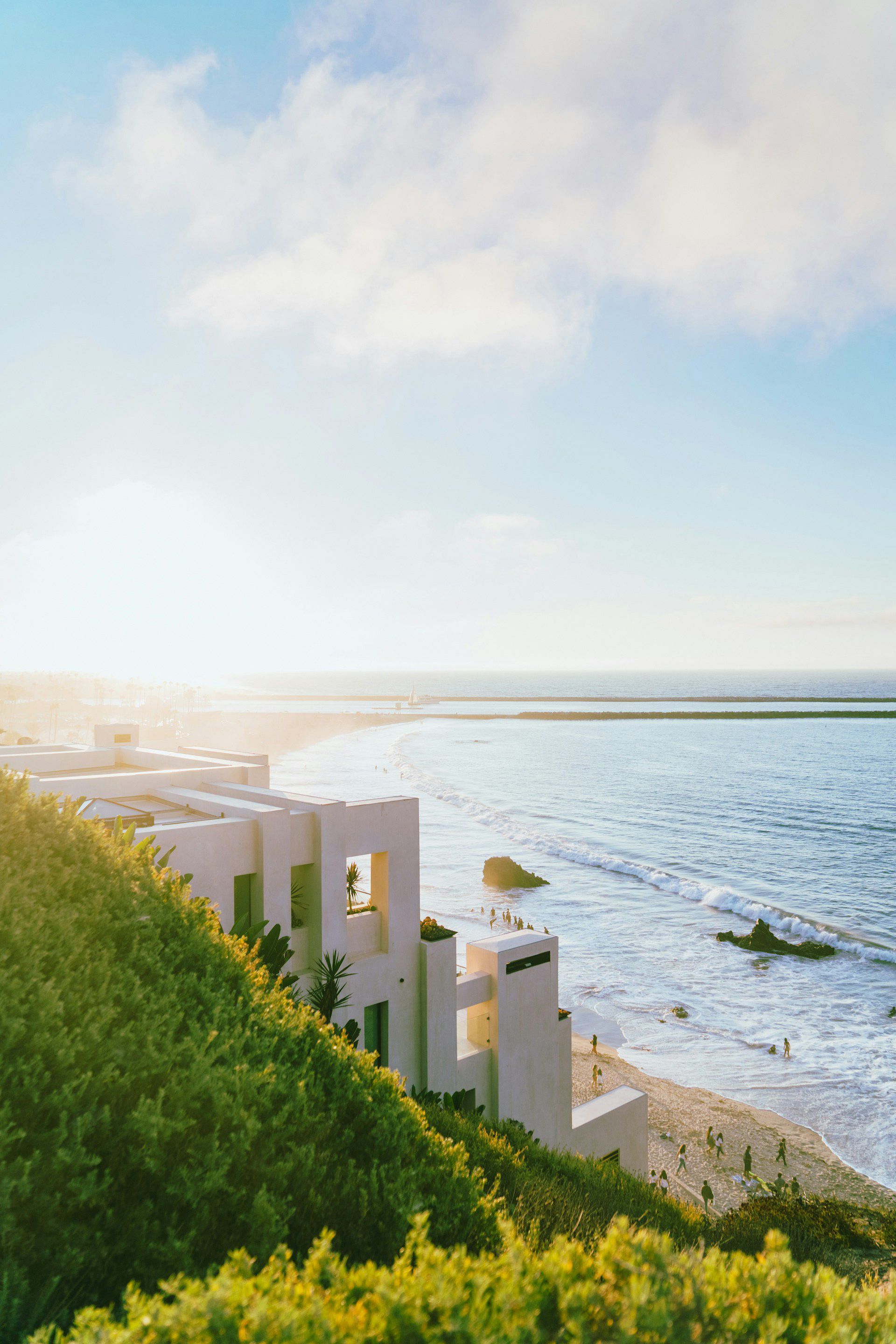 Sunlit seaside view with blue water, a sandy shore, and green hillside with white railing in the foreground