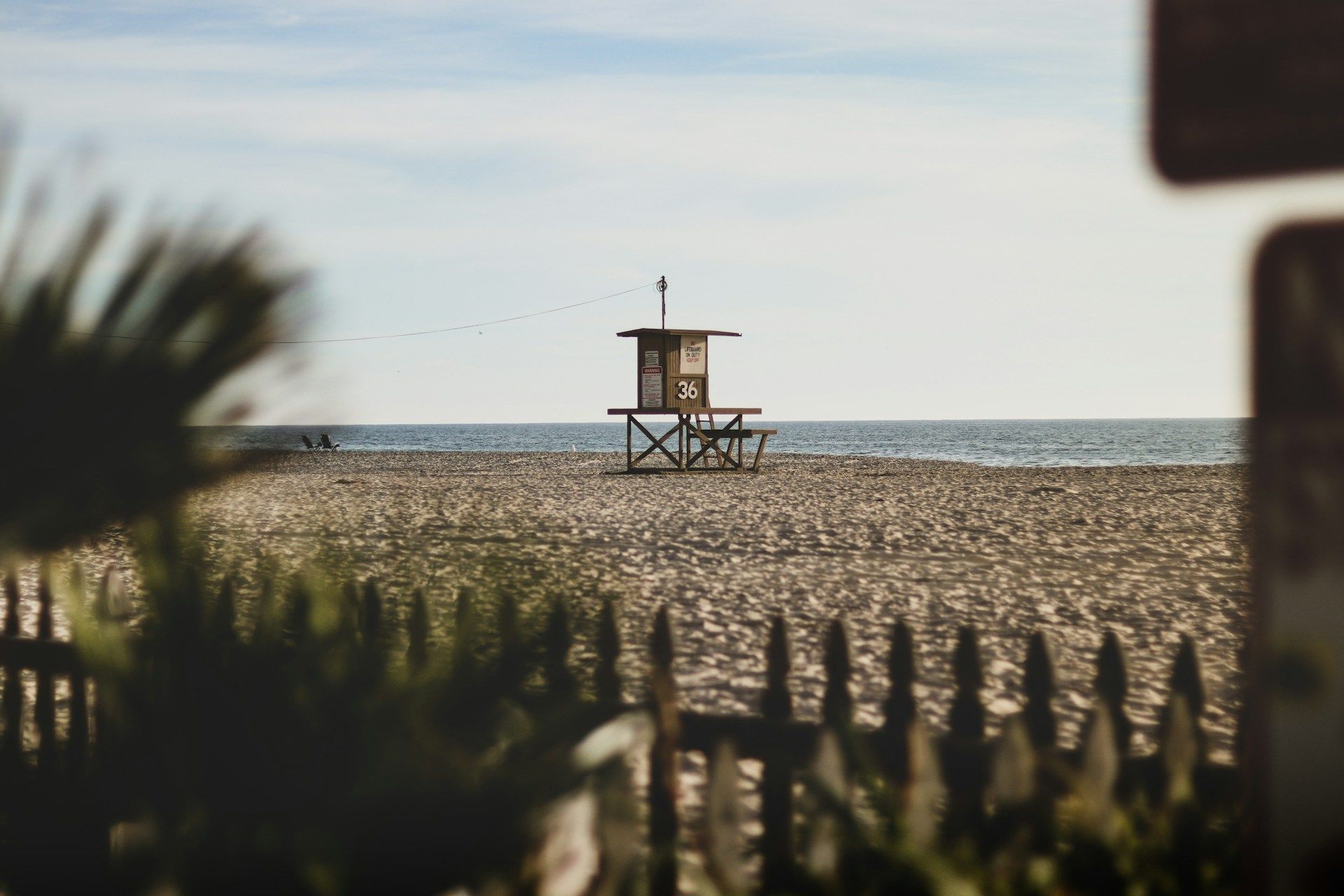 Small lifeguard tower on a sandy beach with ocean horizon, framed by blurred fence and foliage in foreground