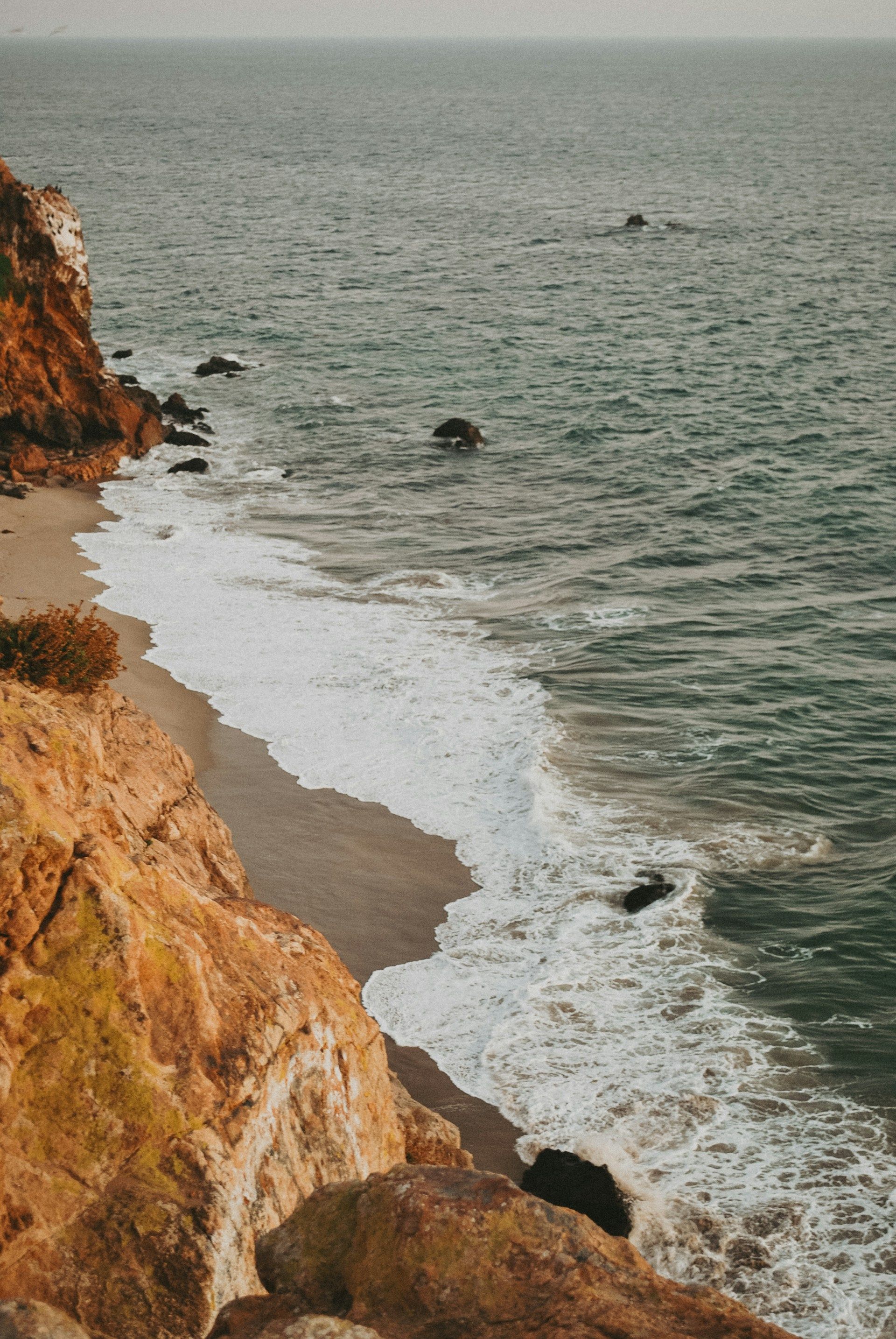 Rocky cliff overlooking a gray, foamy shoreline and calm ocean under an overcast sky