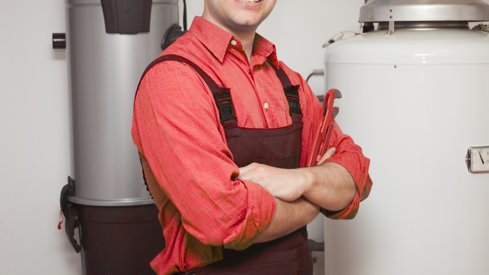 A man in overalls is standing in front of a water heater with his arms crossed.