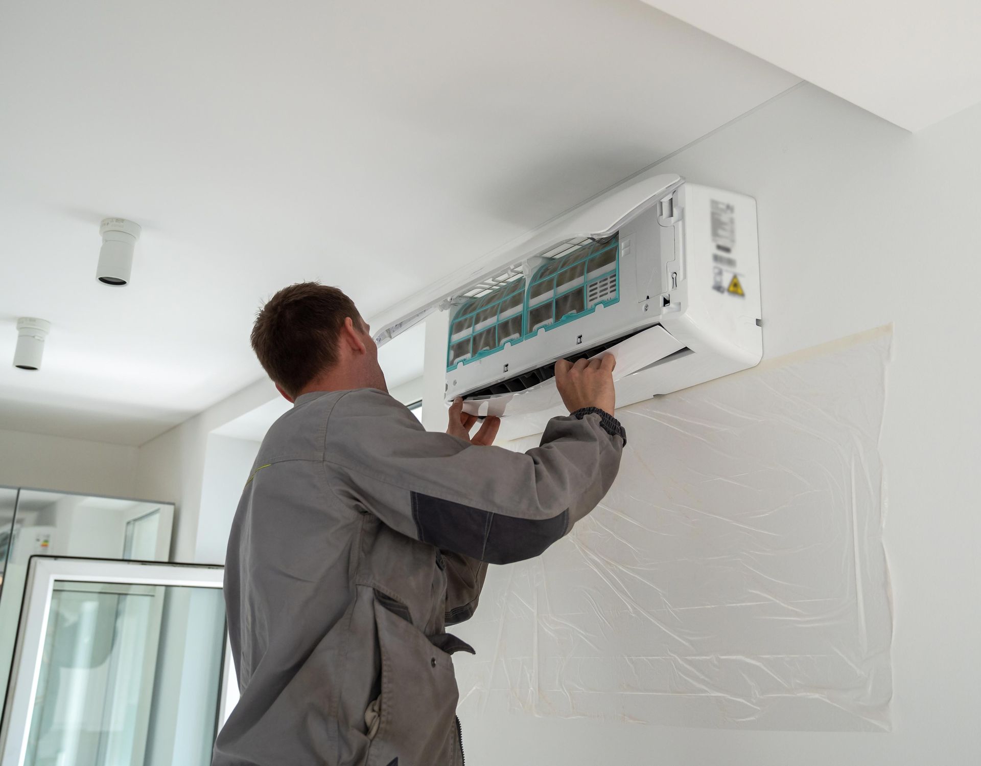 Man installing a white air conditioning unit on a white wall in a room.