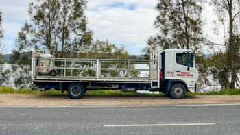 White flatbed truck parked by a roadside, with trees and cloudy sky in the background