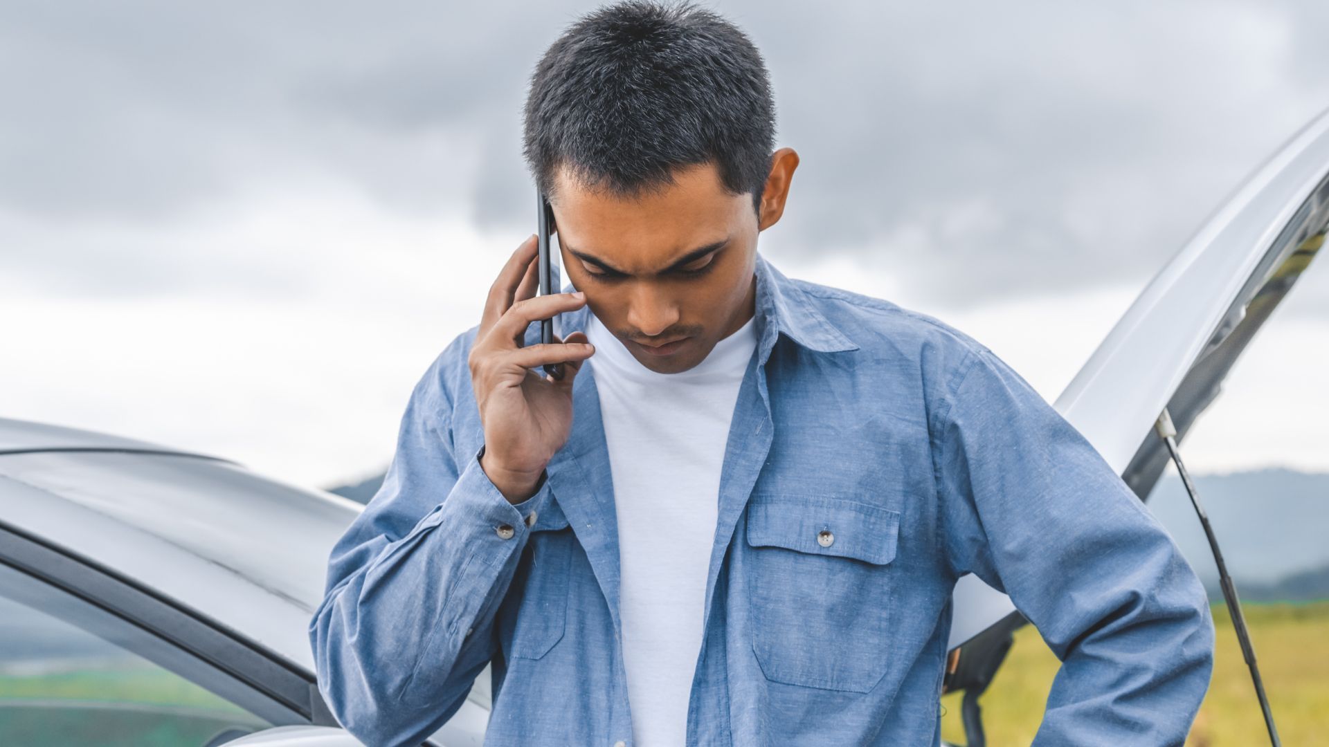 Man on phone beside a car with open hood, looking concerned, overcast sky in background.
