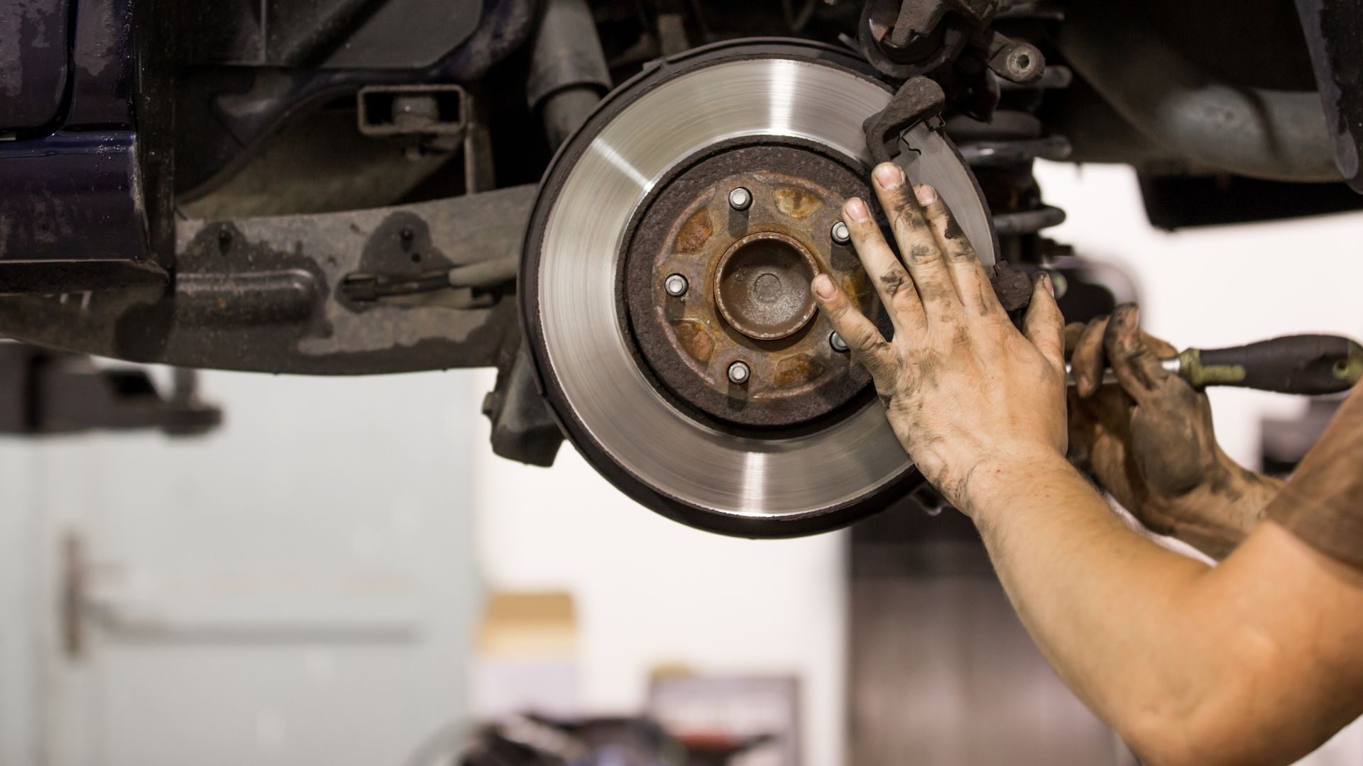 Mechanic working on a car's brake disc and caliper in a garage, hands are dirty.