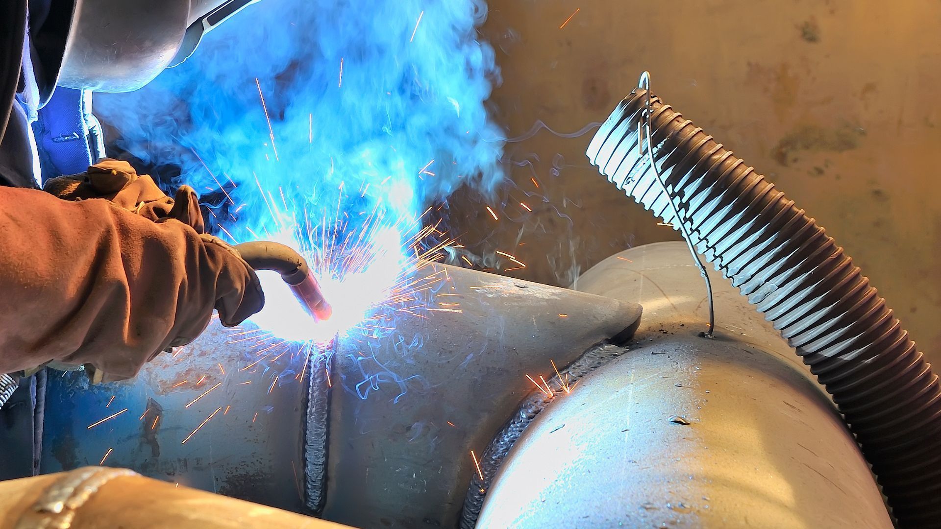 Welder wearing gloves, welding metal pipe, sparks and blue smoke visible.