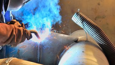 Welder wearing gloves, welding metal pipe, sparks and blue smoke visible.