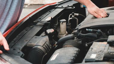 Person opening the hood of a red car, revealing the engine.