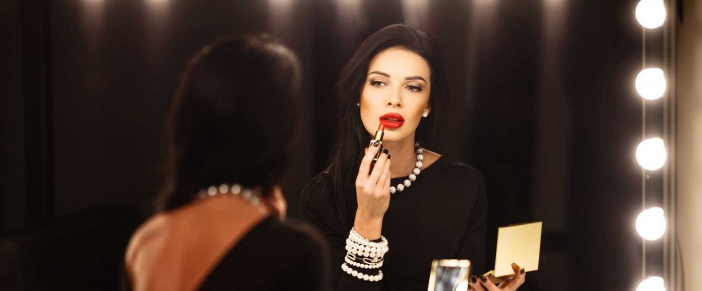 Woman applying red lipstick in front of a vanity mirror with lights, holding a compact mirror.