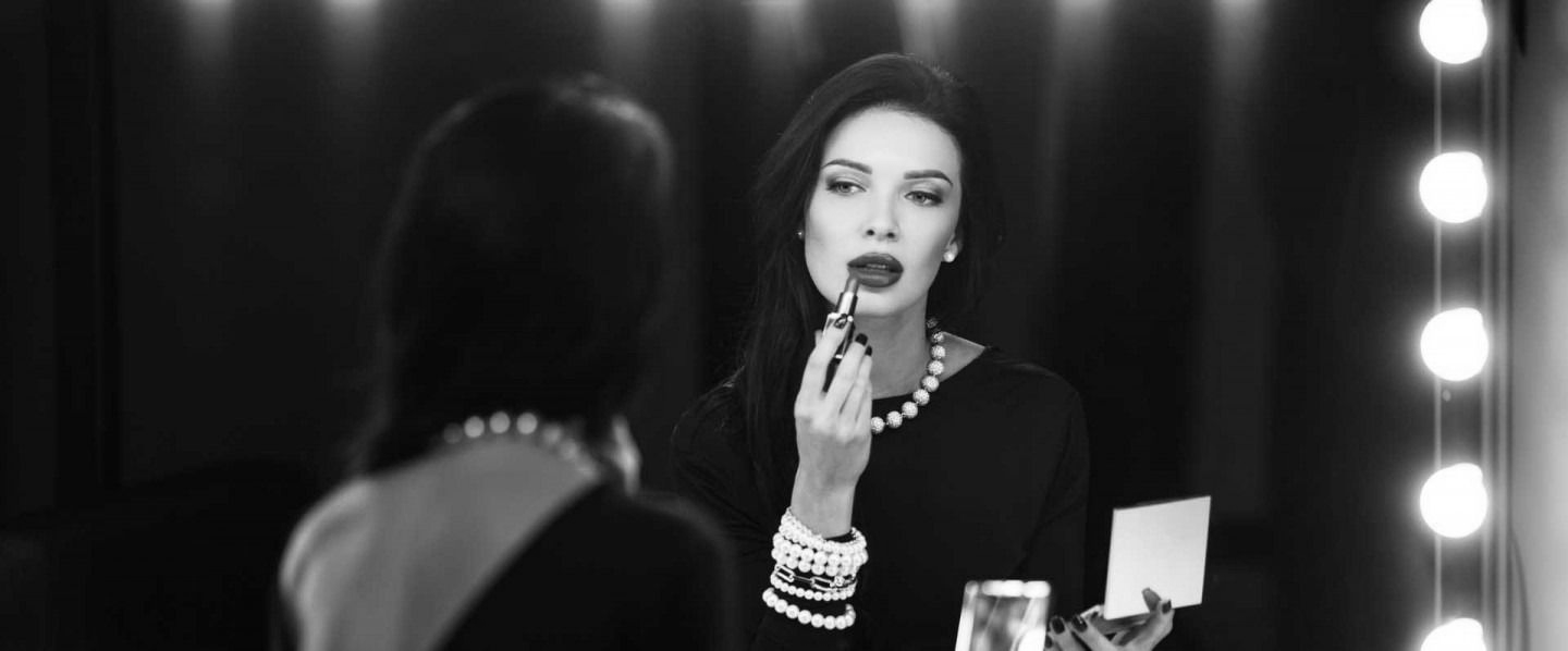 Woman applying lipstick while looking in a mirror, with vanity lights in the background.