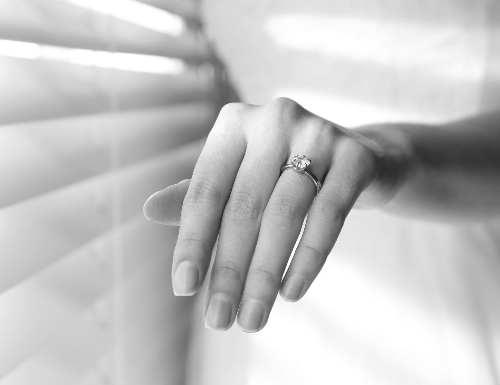Hand with a ring resting against a softly lit window blind, in black and white.