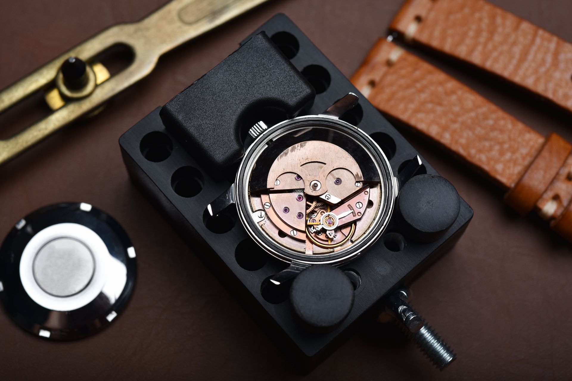 Watch movement on a workbench with tools and brown leather straps nearby