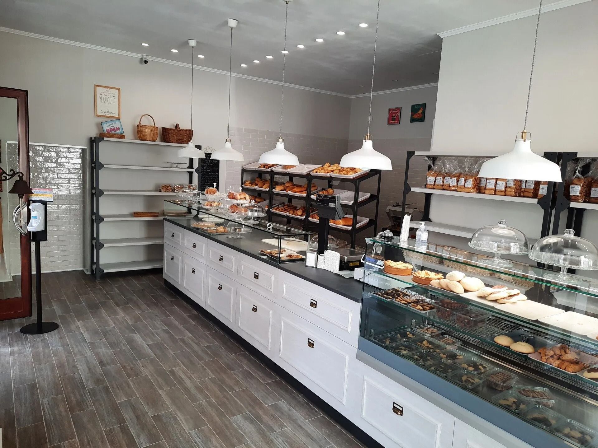 Luminoso bancone da pasticceria con vetrine per dolci, scaffali e lampade a sospensione in un pulito interno grigio.