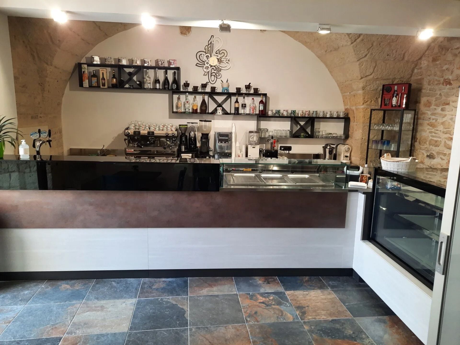 Bancone da bar moderno con macchine da caffè, vetrina e sfondo ad arco in pietra.