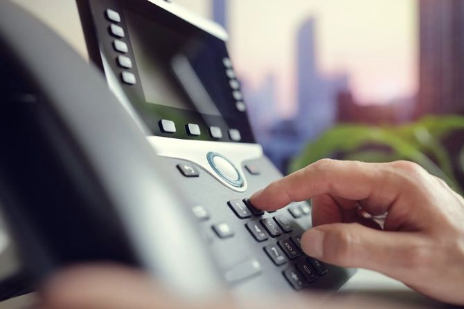 Una mano che preme i tasti di un telefono da ufficio grigio con lo schermo sfocato sullo sfondo.