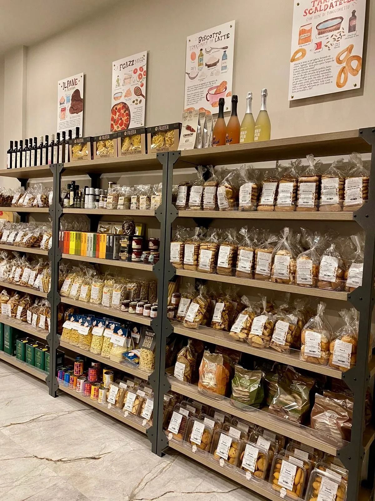 Corridoio del negozio con scaffali pieni di snack confezionati e bottiglie, e poster promozionali appesi in alto.