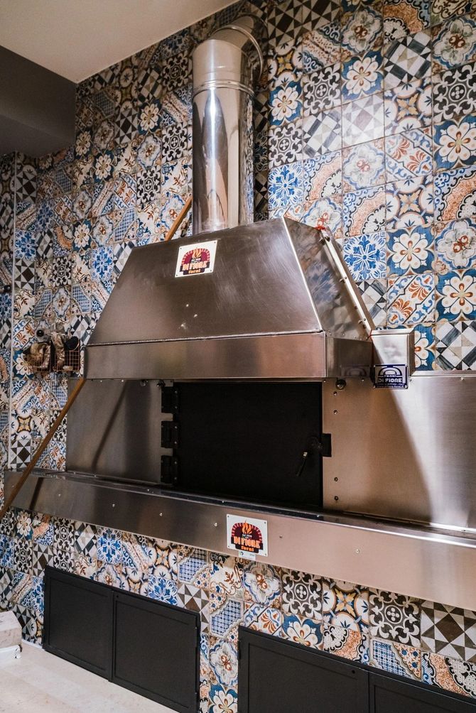 Forno per pizza in acciaio inox in una cucina con piastrelle decorate, con camino e apertura nera.