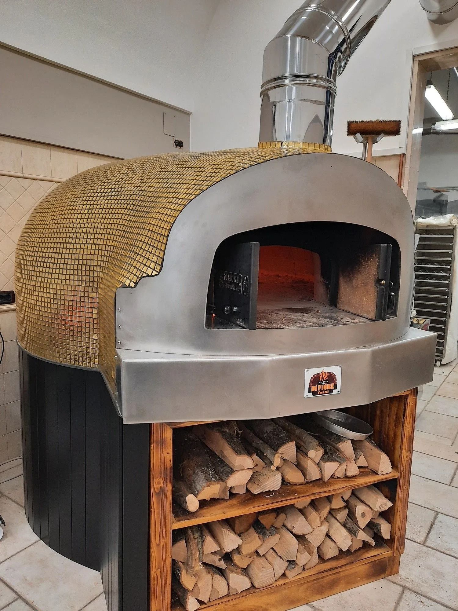 Forno per pizza a legna da esterno con frontale in metallo curvo, camino e legna da ardere accatastata al di sotto.