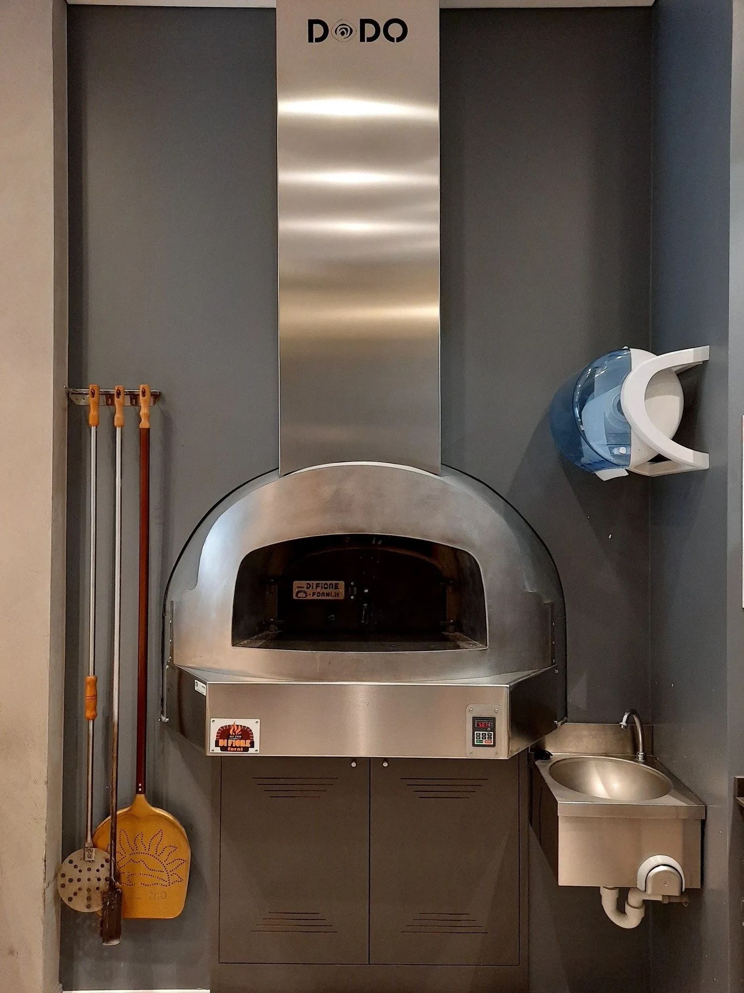 Forno per pizza professionale in una cucina, con utensili per pizza appesi, una pressa per impasto e attrezzature in acciaio inossidabile.