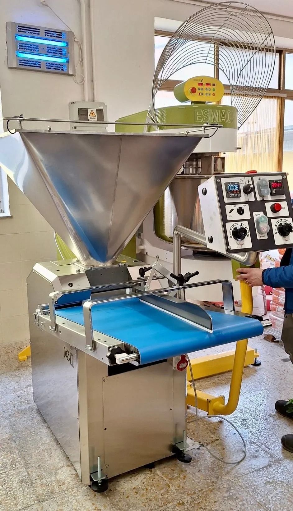 Macchinario industriale con tramoggia in acciaio inox e nastro trasportatore blu in un locale di fabbrica.