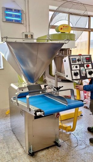 Macchinario industriale con tramoggia in acciaio inox e nastro trasportatore blu in un locale di fabbrica.