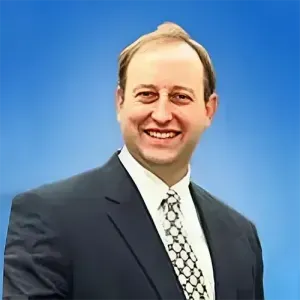 A person wearing a dark suit and patterned tie smiling against a solid blue background.