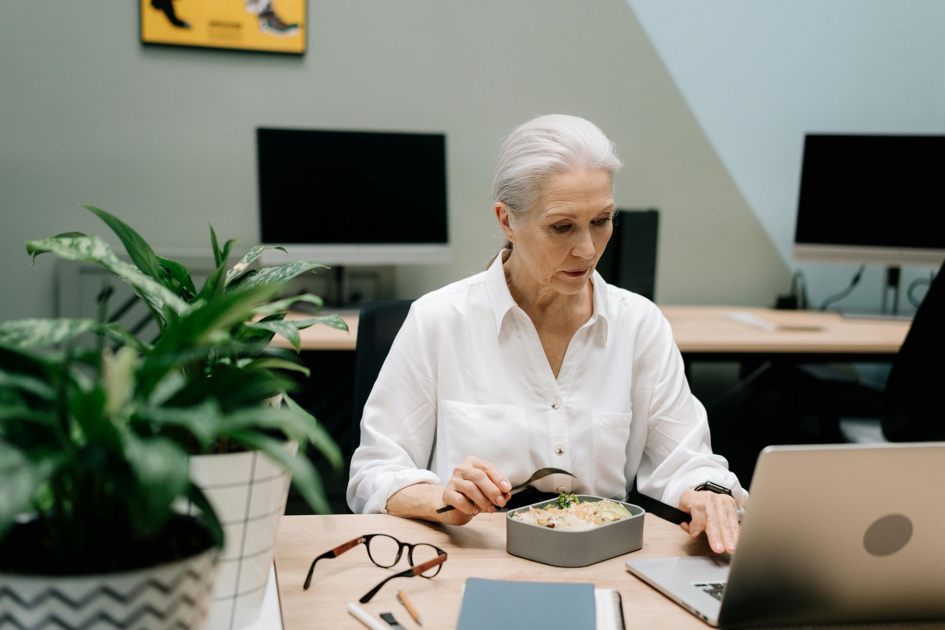 A professional eating a meal at a desk in an office while looking at a laptop computer.