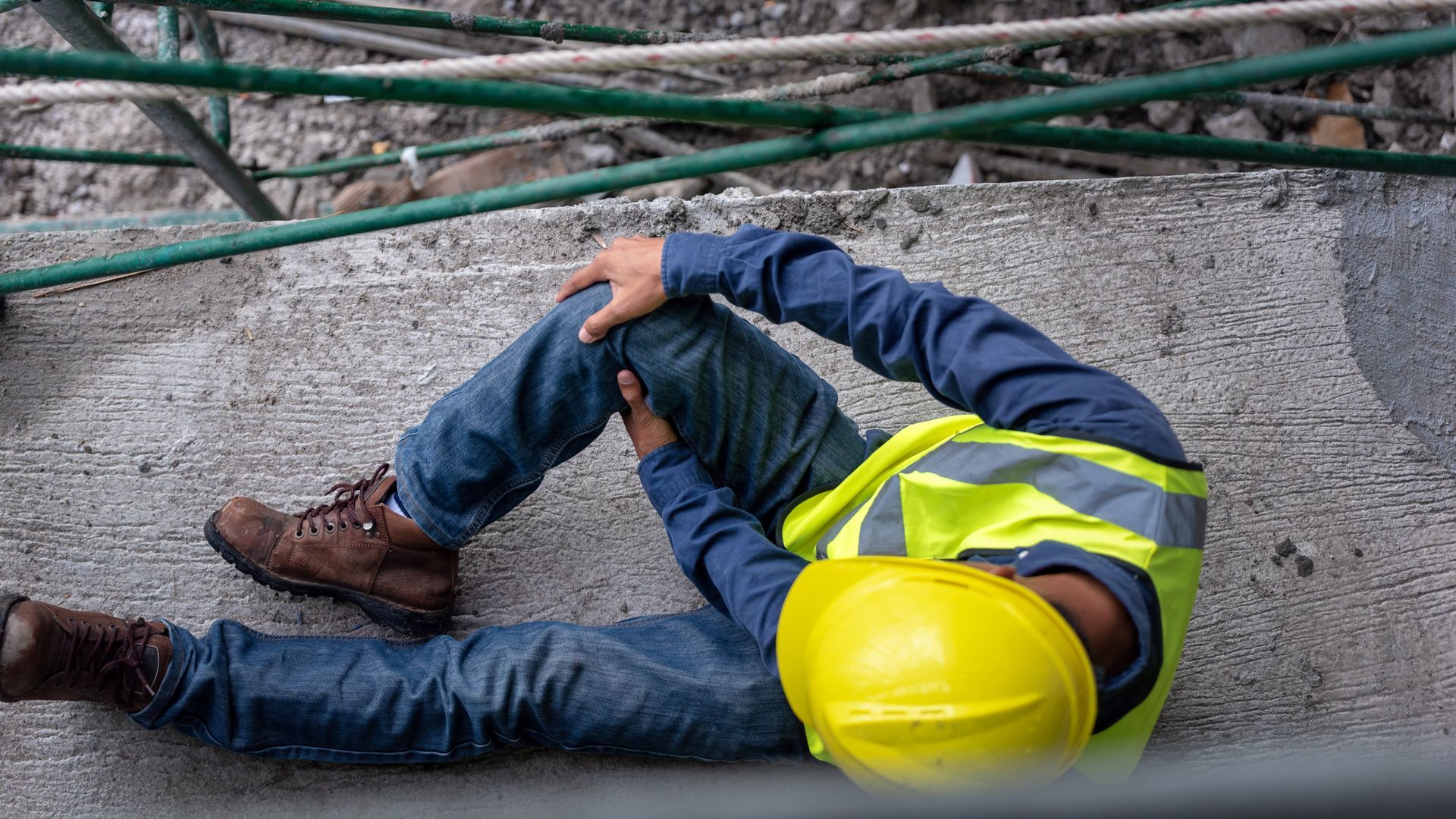 A worker in a yellow hard hat and safety vest sits on concrete, clutching their knee after a fall.