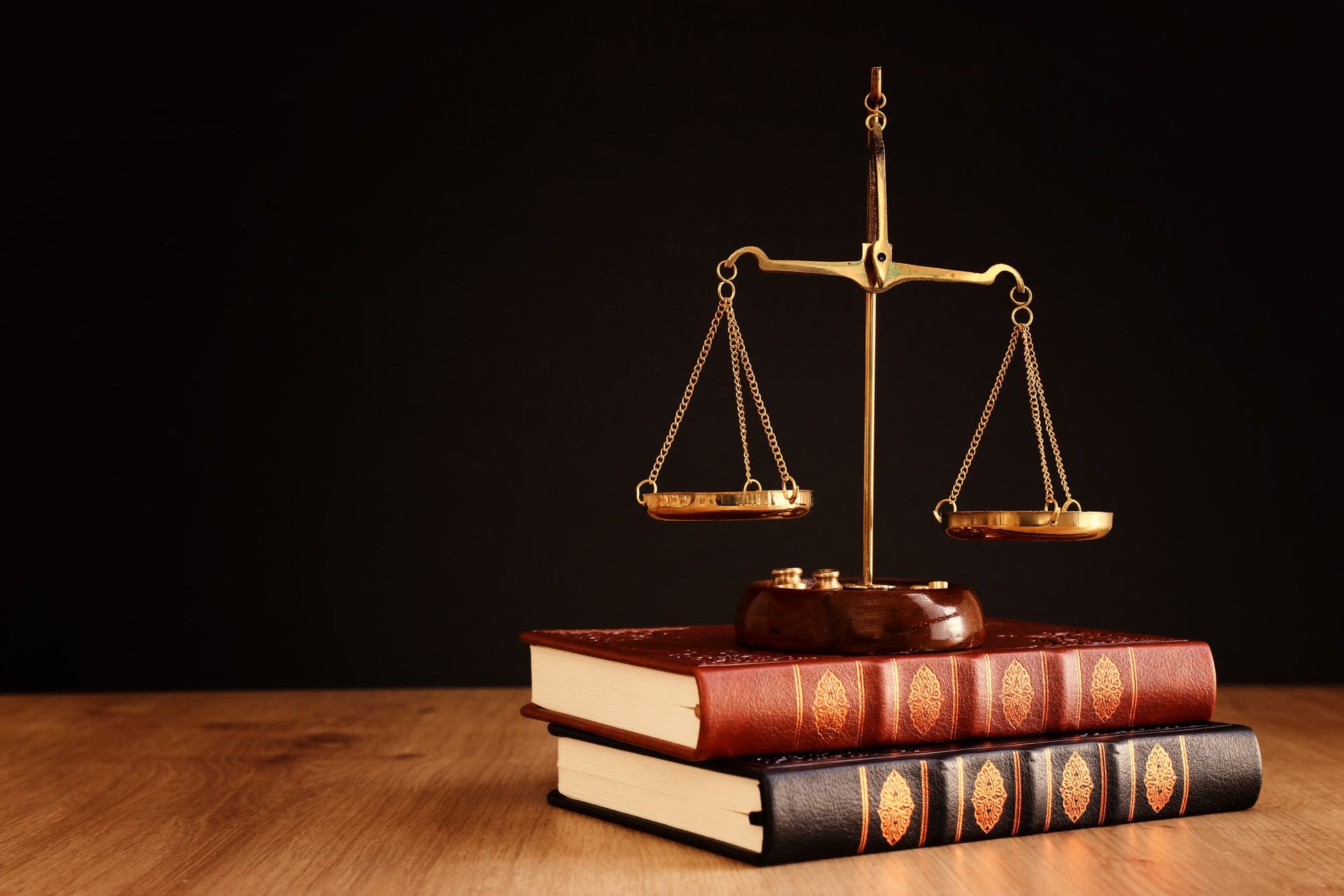 A golden scale of justice sits atop two stacked, vintage-style hardcover books on a wooden surface against a black backdrop.
