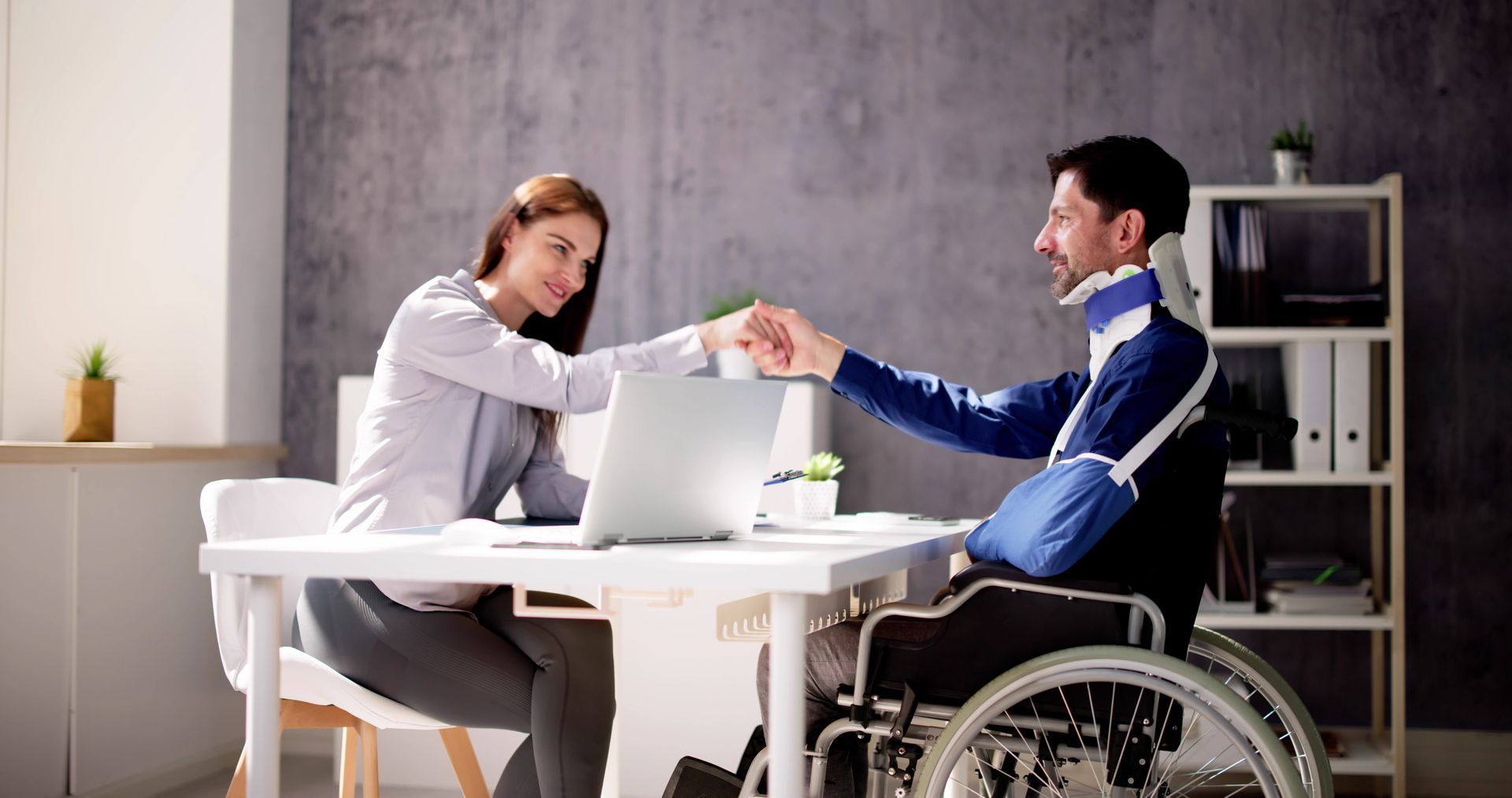 A person in a wheelchair with a neck brace and arm sling fist-bumps a person sitting at a desk with a laptop.