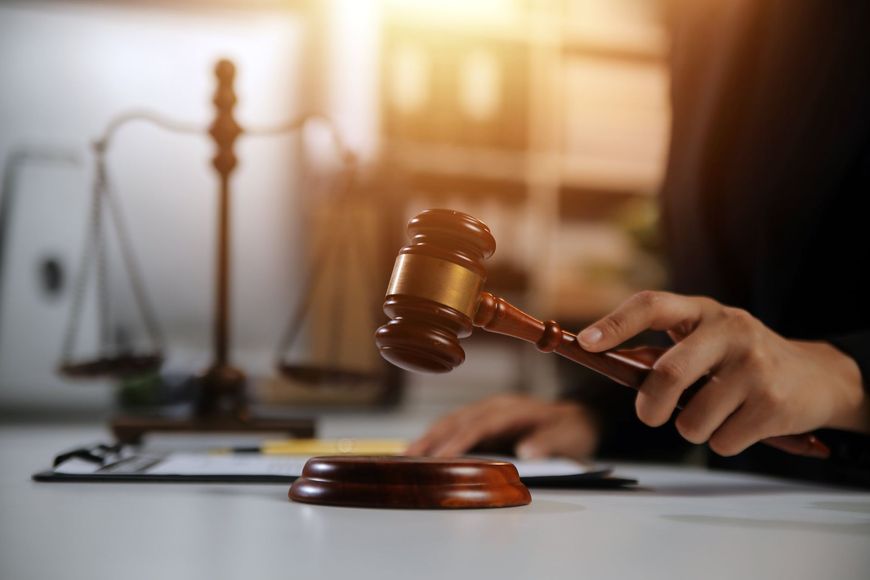 A person holding a wooden gavel over a sound block, with a set of scales of justice in the blurred background.