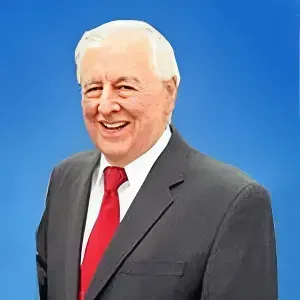 A smiling person wearing a dark suit and red tie stands against a solid blue background.