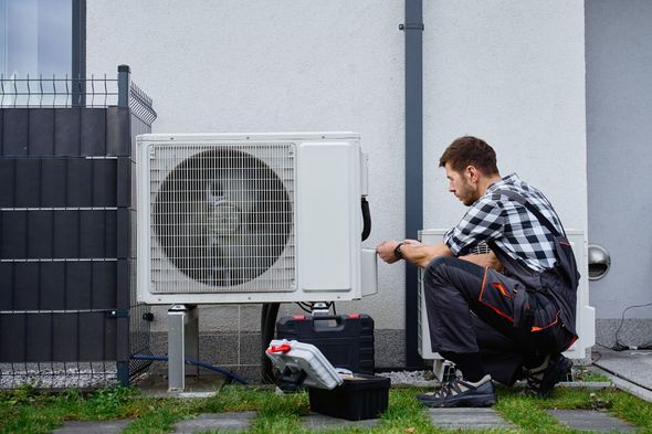 Technician crouches beside an outdoor air conditioner unit, checking wiring by a white wall.