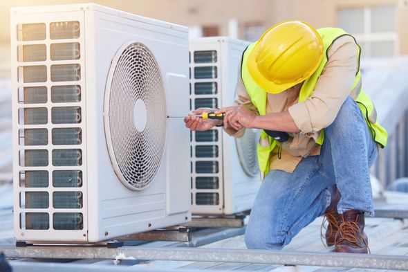 Technician in yellow hard hat kneels beside an outdoor HVAC unit, inspecting it with tools.