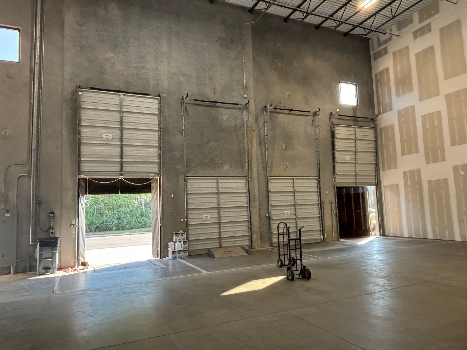 Empty industrial warehouse interior with large metal doors and a concrete floor, lit by daylight.