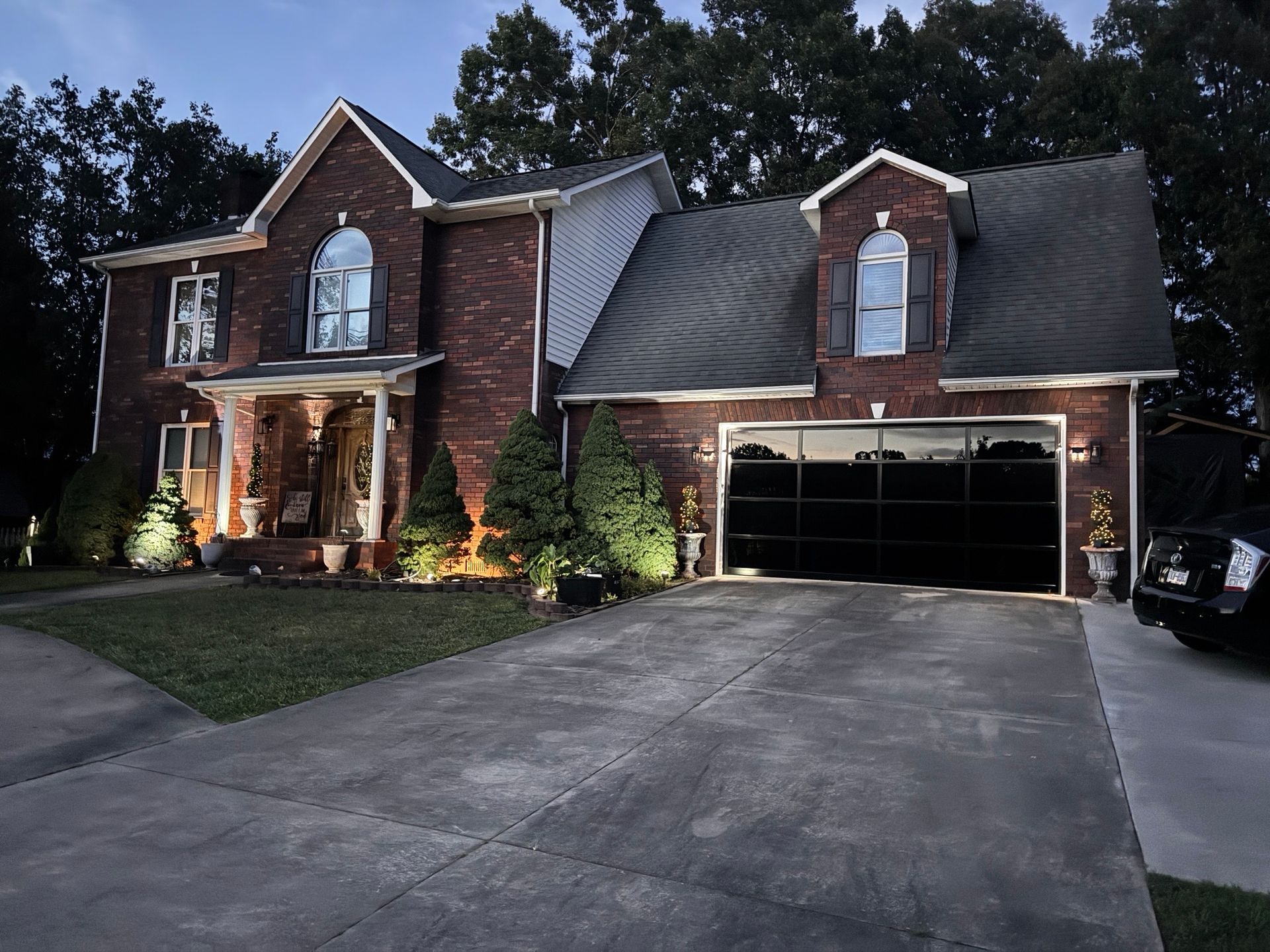 Two-story brick house with lit porch, double garage, and large driveway at dusk