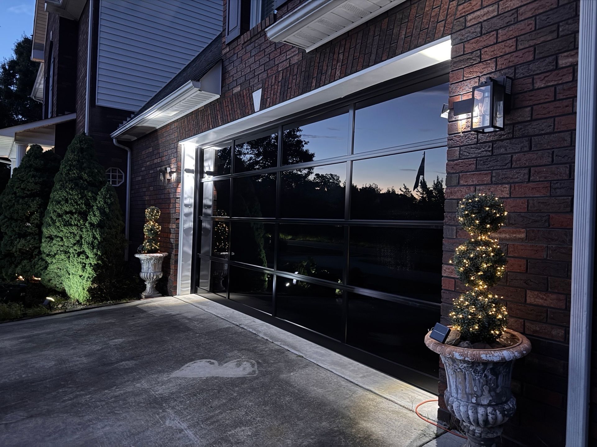 Brick house garage with a glossy black door, lit sconces, and potted evergreen decorations at dusk