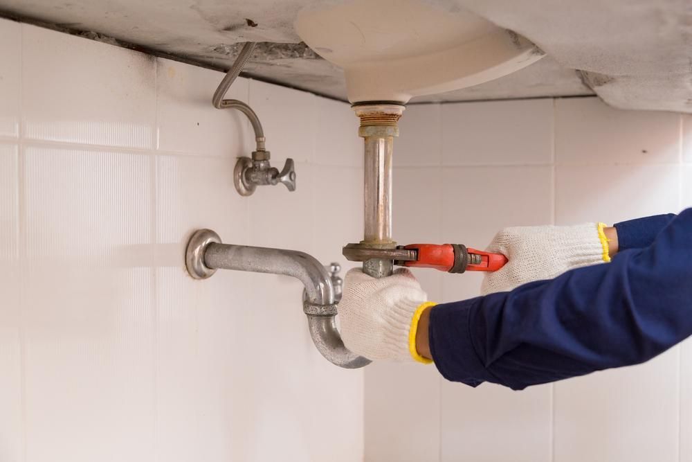 Plumber in Blue Work Shirt and Gloves, Using a Wrench Under a Sink — David McNeill Plumma (Gas Specialist) In Cessnock, NSW