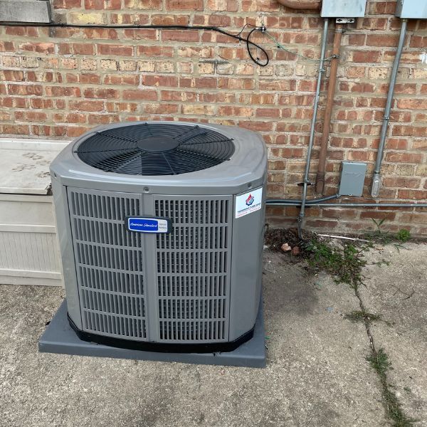 Gray air conditioning unit on a concrete pad, next to a brick wall and electrical conduits.