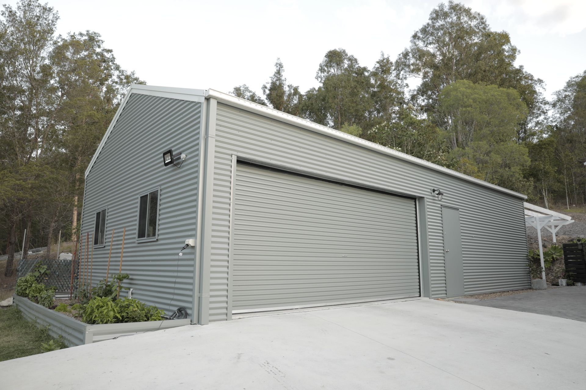 Gray metal garage with a large roller door on a concrete driveway, surrounded by trees.