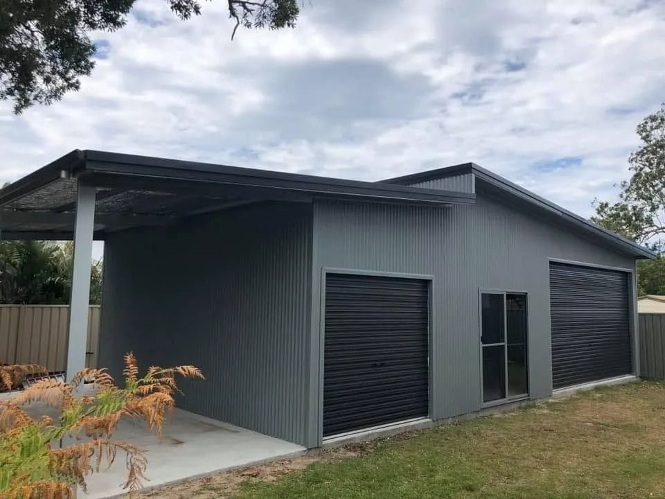 Grey metal garage with black roller doors, a carport, and a side door on a cloudy day.