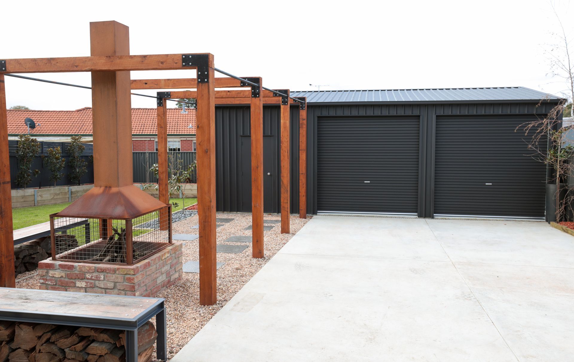 Wooden pergola and fire pit next to a dark grey garage on a concrete driveway.