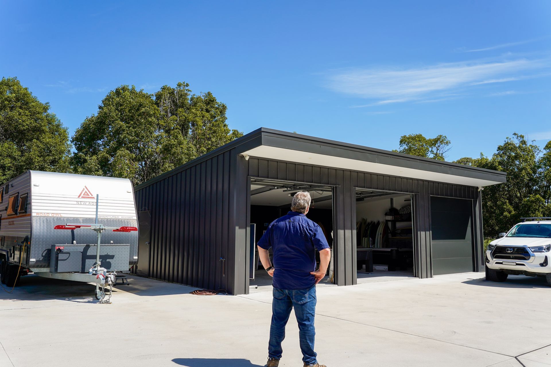 Man standing in front of a dark gray garage, a caravan and truck nearby on a sunny day.