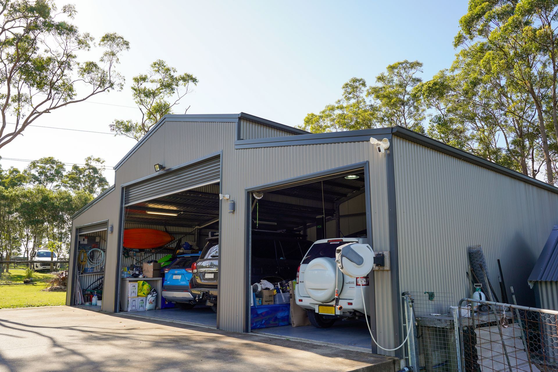 Three-bay metal garage with open doors, housing vehicles and storage. Exterior view with trees in the background.