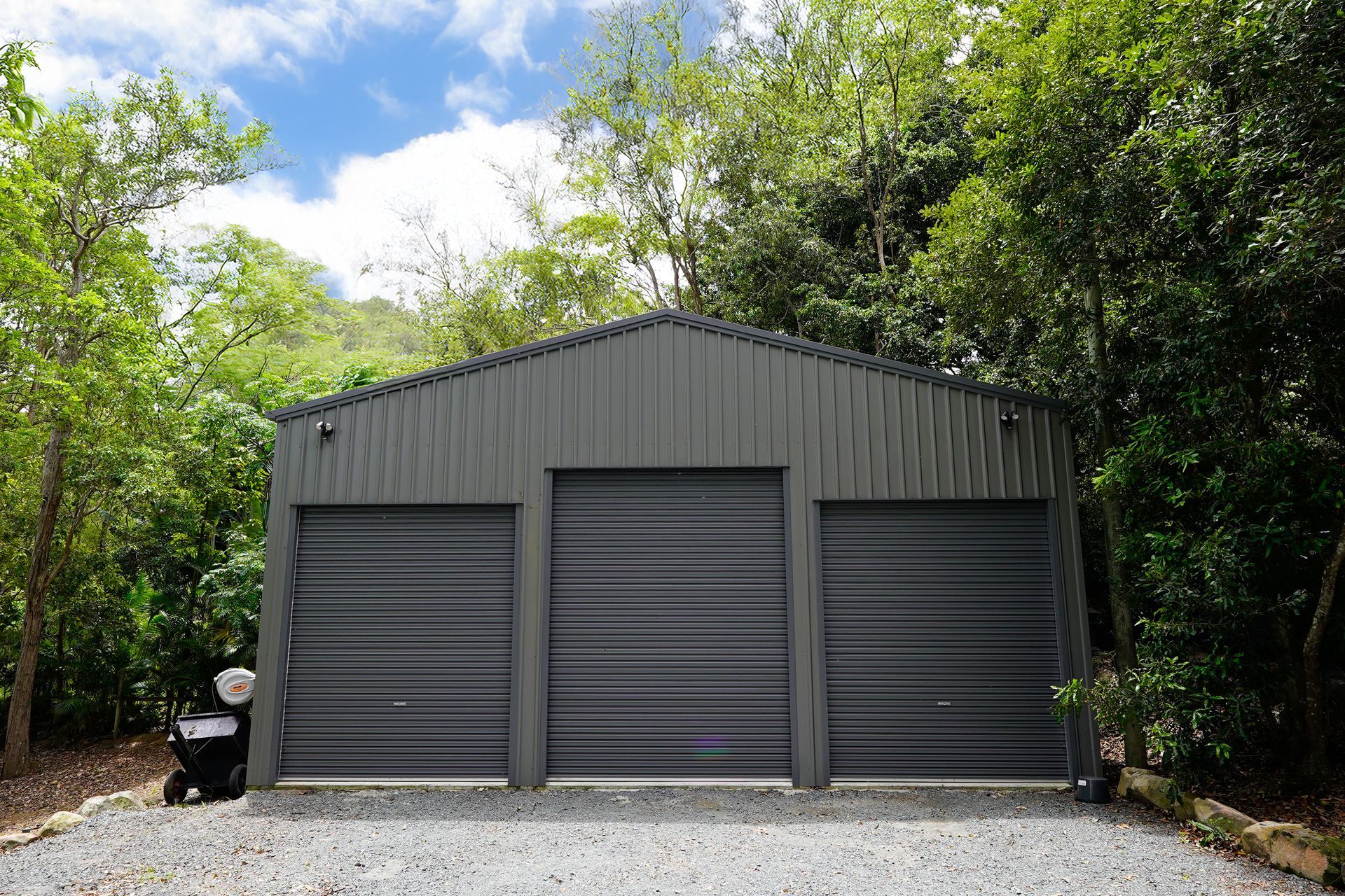 Gray three-bay garage with roller doors, set among trees and gravel driveway.