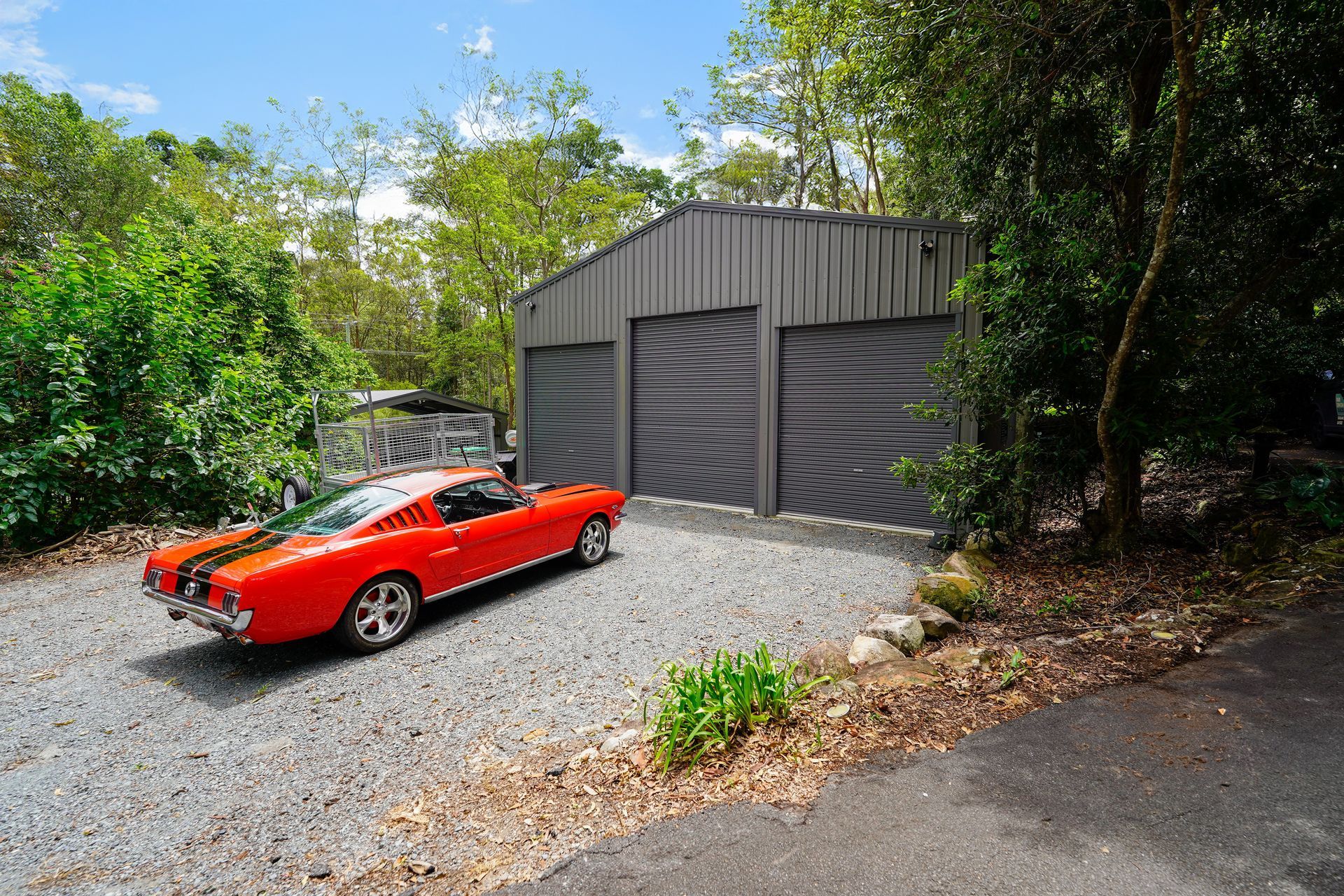 Red classic car parked in front of a gray metal garage on a gravel driveway, surrounded by trees.