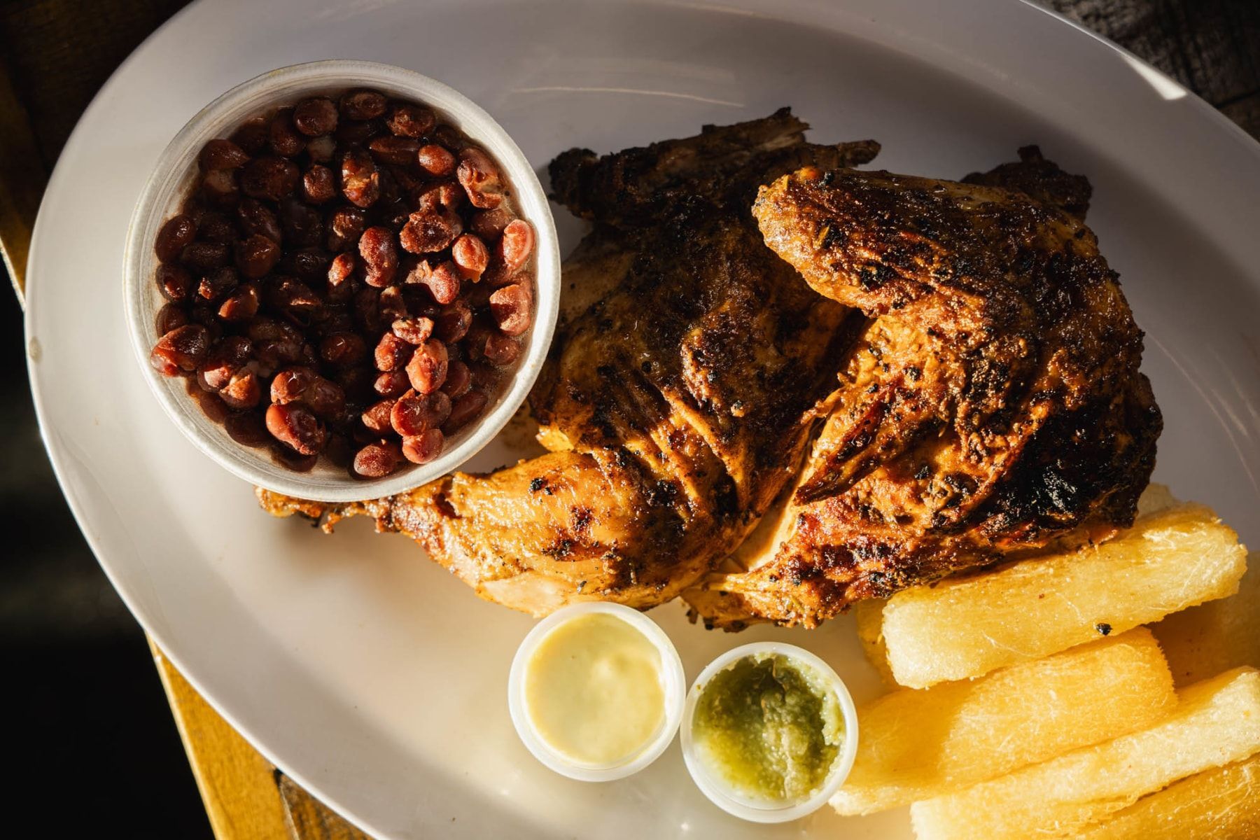 A roasted half chicken served on a white plate with a side of beans, cassava fries, and two small dipping sauces.