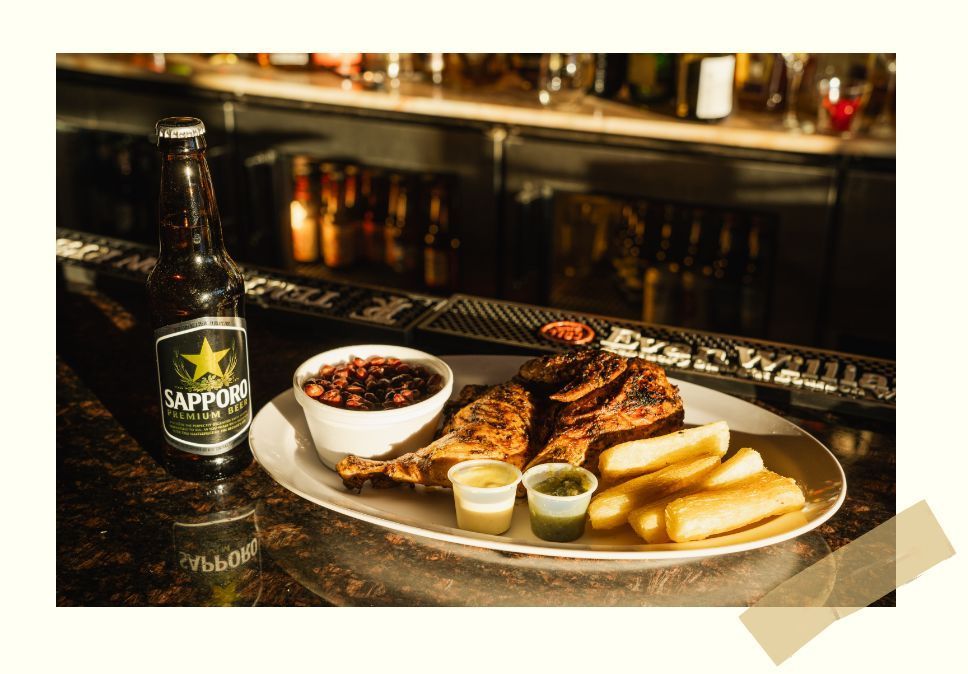 A plate of grilled meat, beans, and fried sides with a bottle of Sapporo beer on a bar counter.