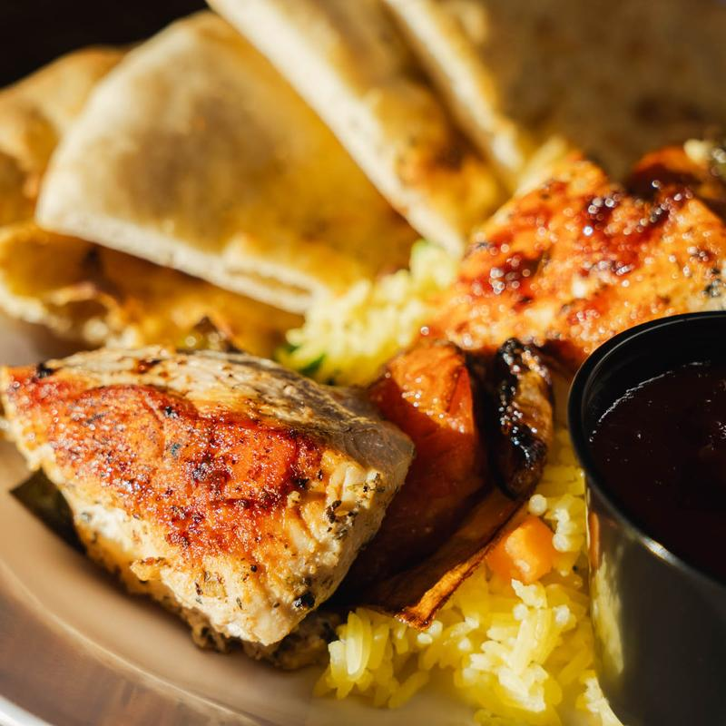Grilled chicken, yellow rice, pita bread, and a small container of dark dipping sauce on a white plate.